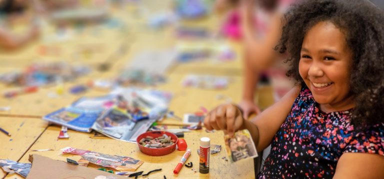 A person holding up some colourful paper grinning, the table to the left of them is covered with crafts and colourful drawings.